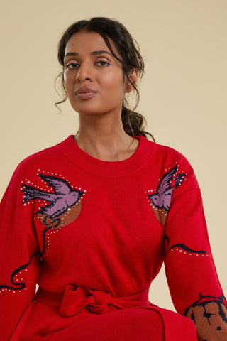 A close up view of a woman modelling a long knitted dress agains a solid beige backdrop. The dress is bright red and belted, and features knitted purple dove designs on the front of each shoulder. Each dove is decorated with crystal detailing, and there are trailing details along the arm and contrasting brown cuffs.
