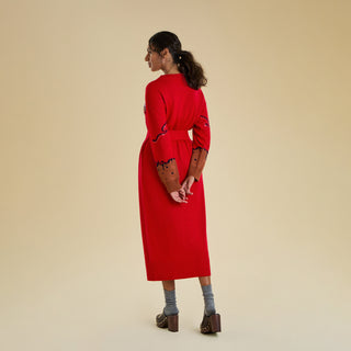 A woman is veiwed from the back modelling a long knitted dress agains a solid beige backdrop. The dress is bright red and belted, and there are trailing details along the arm and contrasting brown cuffs.