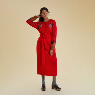 A woman models a long knitted dress agains a solid beige backdrop. The dress is bright red and belted, and freatures knitted purple dove designs on the front of each shoulder. Each dove is decorated with crystal detailing, and there are trailing details along the arm and contrasting brown cuffs.
