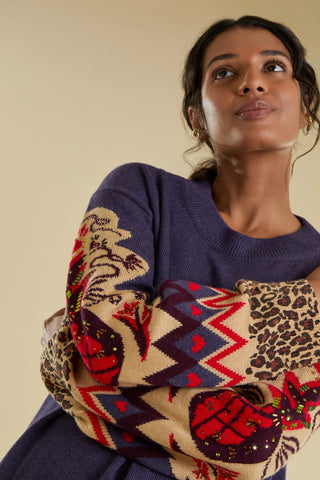 A woman is modelling a knitted jumper against a solid beige backdrop. She has her arms crossed in front of her to display intricate designed sleeves. The jumper is a purple colour and features a large  knitted pineapple on the sleeve with crystal embellishment, knitted zig zag and heart banding, and an animal print cuff.