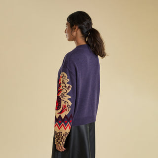 A woman is modelling a knitted jumper against a solid beige backdrop. The jumper is a purple colour and features a large  knitted pineapple on the sleeve with crystal embellishment, knitted zig zag and heart banding, and an animal print cuff.