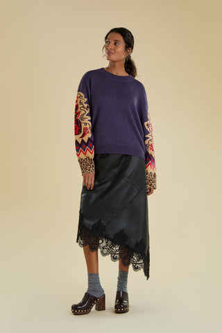 A woman is modelling a knitted jumper against a solid beige backdrop. The jumper is a purple colour and features a large  knitted pineapple on the sleeve with crystal embellishment, knitted zig zag and heart banding, and an animal print cuff.