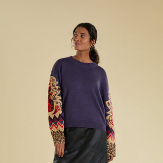 A woman is modelling a knitted jumper against a solid beige backdrop. The jumper is a purple colour and features a large  knitted pineapple on the sleeve with crystal embellishment, knitted zig zag and heart banding, and an animal print cuff.