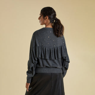 A woman is viewed from behind modelling a grey knitted jumper. The sweater has sparkly crystal decoration and features an elegant ruffle that forms a V shape in the centre of the back and trails down the arms.