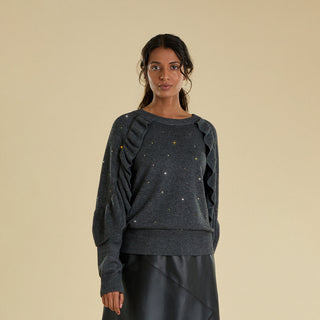 A woman modeling a dark gray knit sweater with delicate multi coloured crystal embellishments, and elegant ruffle detail down the sleeve, paired with a black skirt, against a solid beige background.