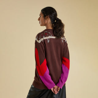 A woman shown from the back wearing a brown knit sweater with red and purple colour-blocked sleeves and crystal embellishments on the shoulders, set against a solid beige background.