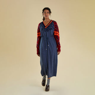 A woman modeling a long knitted dress against a solid beige studio backdrop. The dress is a denim blue colour and has contrasting dark red raglan sleeves featuring sporty stripes and swirl detail decorated with gold studs. The chest has tonal animal print motifs that fan out from beneath a deep V neckline.