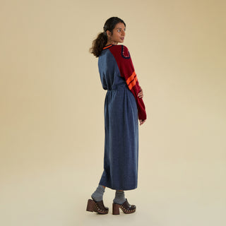 A woman modeling a long knitted dress against a solid beige studio backdrop. The dress is a denim blue colour and has contrasting dark red raglan sleeves featuring sporty stripes and swirl detail decorated with gold studs.