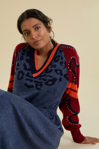 A woman is sitting down modeling a long knitted dress against a solid beige studio backdrop. The dress is a denim blue colour and has contrasting dark red raglan sleeves featuring sporty stripes and swirl detail decorated with gold studs. The chest has tonal animal print motifs that fan out from beneath a deep V neckline.