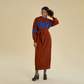 A woman models a long knitted dress against a solid beige background. The belted dress is an earthy brown colour with an electric blue zig zag pattern on the sleeve and chest. 
