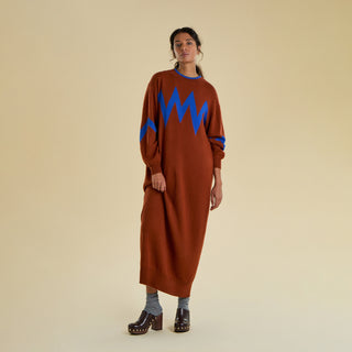 A woman models a long knitted dress against a solid beige background. The dress is an earthy brown colour with an electric blue zig zag pattern on the sleeve and chest. 