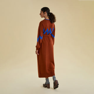 A rear view of a woman modeling a long knitted dress against a solid beige background. The belted dress is an earthy brown colour with an electric blue zig zag pattern on the sleeve and chest. 