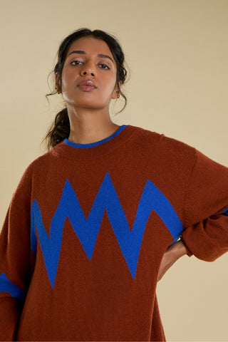 A woman wearing a rust-brown knit dress with a bold blue zigzag pattern across the chest and blue trim at the collar, posed against a solid beige background.